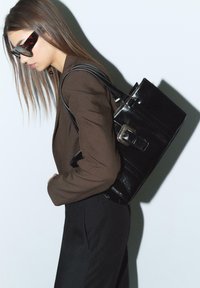 Sac fourre-tout noir brillant au design structuré, avec un détail de boucle en argent et deux bretelles, sur fond uni.