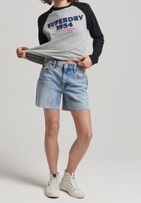 Graues und schwarzes Langarm-T-Shirt mit dem Aufdruck "SUPERDRY 1954", kombiniert mit ausgefransten blauen Denim-Shorts und weißen Sneakers.