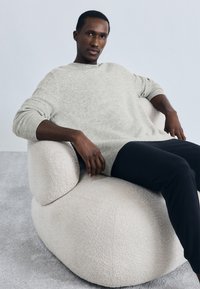 Άνδρας που φοράει ανοιχτό γκρι πουλόβερ και μαύρο παντελόνι καθισμένος χαλαρά σε μια καρέκλα από μπουκλέ εκρού μπροστά σε ένα απλό ανοιχτόχρωμο φόντο.