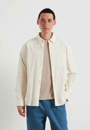 Camicia beige chiaro con bottoni e taschino sul petto, indossata sopra una t-shirt color sabbia, abbinata a pantaloni blu. Tessuto liscio, design casual.