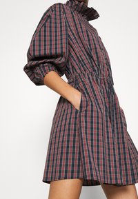 Rutiga klänningen har hög krage, puffärmar, cinchad midja och sidofickor. Tyget är marinblått med röda och vita ränder.