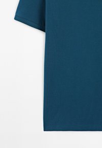 Rukáv a boční část jednoduchého tmavě modrozeleného trička s krátkým rukávem na bílém pozadí.