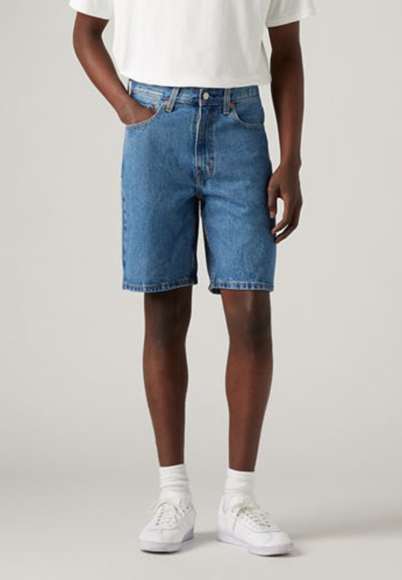 Denimshorts i mellomblå, avslappet passform, med fem lommer og beltehemper. Brukt med hvite joggesko og ankelsokker.