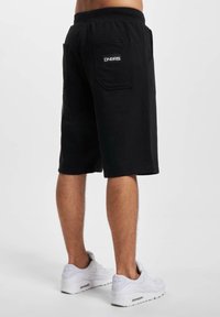 Black cotton shorts with an elastic waistband and back pocket, featuring a small logo patch. The hem is straight and ends just above the knee.