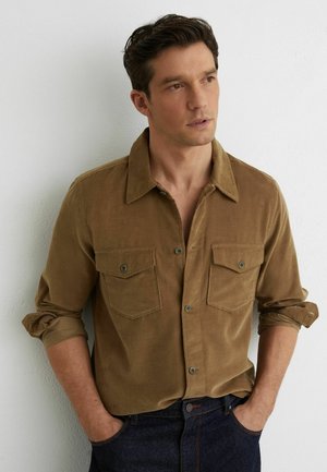 Mężczyzna o ciemnych włosach, ubrany w brązową koszulę z podwiniętymi rękawami i ciemne jeansy, stoi opierając się o białą ścianę, trzymając ręce w kieszeniach.