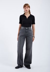 Schwarzes geripptes Poloshirt mit Kragen, kombiniert mit hoch taillierten, weit geschnittenen, dunkelgrauen Jeans. Das Modell trägt schwarze Schuhe; minimalistisches Design.