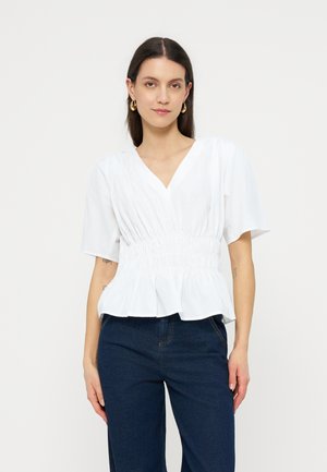 Frau mit dunklen Haaren, die goldene Creolen, eine weiße V-Ausschnitt-Bluse mit Taillierung und dunkelblaue Jeans trägt, steht vor einem hellen Hintergrund.