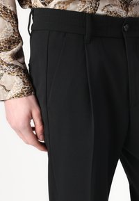 Zwarte op maat gemaakte broek met plooien, met een vlakke voorkant, knoopsluiting en elastische tailleband. Gladde stof met een strakke afwerking.