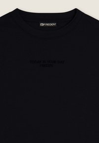 Camiseta de algodón negra con cuello redondo y texto en relieve que dice "HOY ES TU DÍA FREDDY" en el centro. Textura suave.