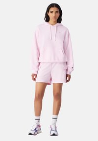 Felpa con cappuccio di un rosa chiaro, dotata di tasca frontale, abbinata a pantaloni corti coordinati. Sneakers bianche da ginnastica con dettagli viola e calze a righe.
