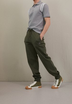 Mand iført grå polo shirt, olivengrønne joggebukser og grønne høje sneakers med hvide snørebånd, stående mod en ensfarvet beige væg.