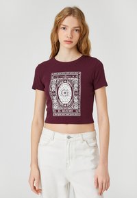 Mørkerød kortærmet t-shirt lavet af ribbet stof, der har et hvidt grafisk mønster med teksten "EL MUNDO" og et øje-motiv.
