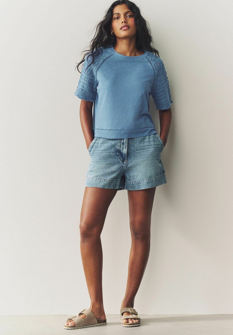 Light blue cotton top with textured sleeves, paired with faded denim shorts and taupe slip-on sandals featuring a decorative buckle.
