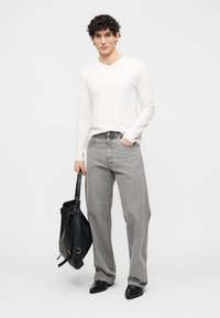 Νέος άνδρας με λευκό μακρυμάνικο πουκάμισο, άνετο γκρι τζιν, μαύρα παπούτσια, κρατώντας ένα μαύρο σακίδιο στο πλάι, μπροστά σε λευκό φόντο.