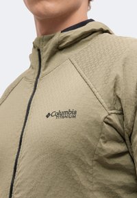 Άτομο που φοράει ανοιχτοπράσινο μπουφάν Columbia Titanium με υφή υφάσματος και μαύρο φερμουάρ σε απλό φόντο.