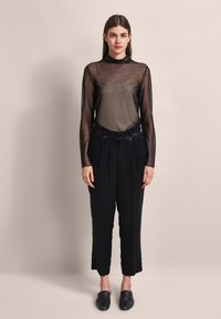 Schwarzes, langärmliges, durchsichtiges Mesh-Oberteil über einer schwarzen, high-waisted, maßgeschneiderten Hose mit geradem Bein. Getragen mit flachen, schwarzen Slippern.