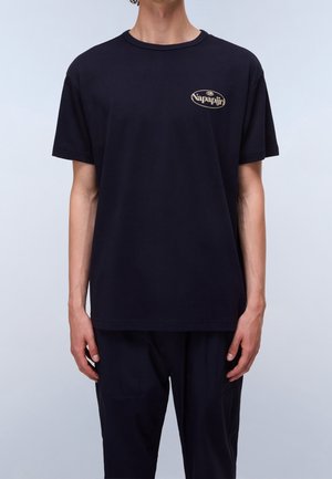 Personne portant un T-shirt noir uni à manches courtes avec un petit logo Napapijri beige sur la poitrine gauche.