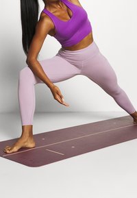 Fialový sportovní top a světle fialové legíny na osobě v bojovém postoji, stojící na tmavě fialové jógové podložce s liniovými značkami.