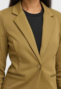 Khaki blazer s upraveným střihem, tvarovanými klopami, uzávěrem na jeden knoflík a dvěma předními kapsami, nošený přes černou košili.
