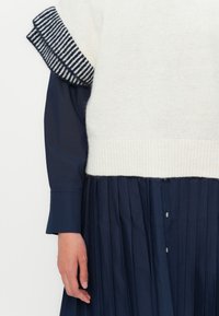 Weißer Strickpullover mit gestreiften marineblauen Akzenten an den Ärmeln, kombiniert mit einem dunkelmarineblauen plissierten Kleid mit Knopfdetails und einer glatten Textur.