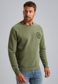 Olive green sweatshirt with a crew neckline, ribbed cuffs, and a textured patch logo on the chest. Fabric has a soft, smooth finish.