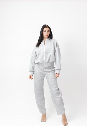 Frau steht und trägt einen hellgrauen Reißverschluss-Sweatshirt und passende Jogginghose mit klaren Sandaletten mit hohem Absatz vor weißem Hintergrund.