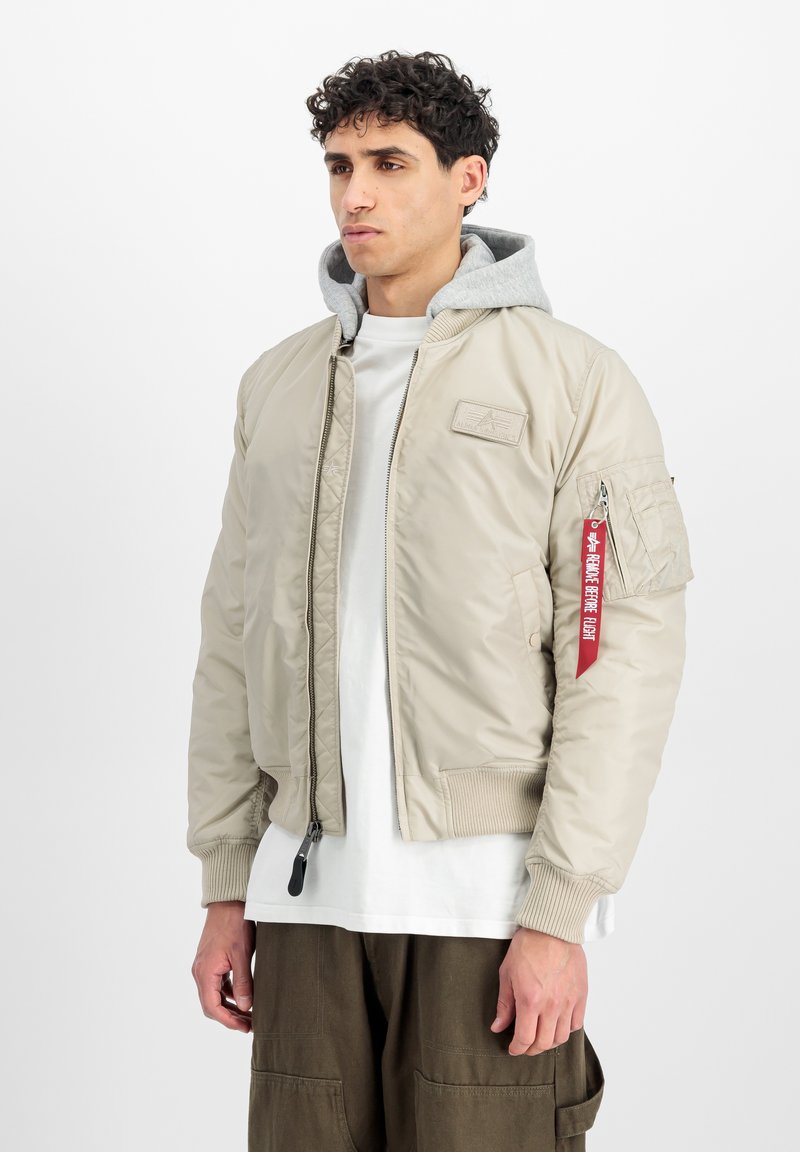 Beige bomber jacket with ribbed cuffs, zip front, and a grey hood. Features a chest patch and side pocket with red accent tag.