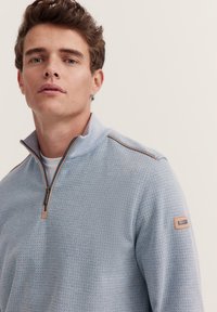 Hellblauer Halb-Zip-Pullover aus strukturiertem Strickstoff, mit kontrastierendem dunklen Reißverschluss und einem lederähnlichen Patch am linken Ärmel.