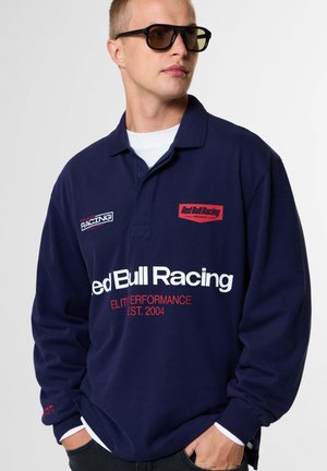 Uomo con occhiali da sole e polo a maniche lunghe Red Bull Racing blu navy con loghi bianchi e rossi, in piedi con le mani in tasca.