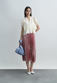 Γυναίκα που φοράει λευκό μπλουζάκι από δαντέλα με λουλούδια, φούστα σε πλισέ σε απαλό ροζ, λευκά μυτερά παπούτσια, κρατώντας μια απαλής μπλε απόχρωσης τσάντα, στέκεται μπροστά σε γκρίζο φόντο.