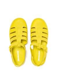 Las sandalias de plástico amarillas cuentan con un diseño de correas entrelazadas, una hebilla ajustable en el tobillo y una plantilla texturizada. Tienen una superficie lisa y costados abiertos.