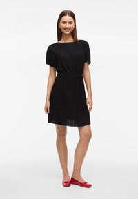 Vestido plissado preto com mangas curtas e cintura ajustada, combinado com sapatos planos vermelhos. Tecido suave, comprimento até o joelho.