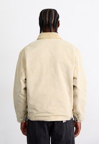 Veste en toile beige avec un col, des coutures visibles et des boutons-pression à l'ourlet. Portée sur un pantalon noir texturé.