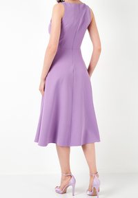 Femme portant une robe midi sans manches lavande avec fermeture éclair au dos et des sandales à talons assorties, se tenant contre un fond blanc.