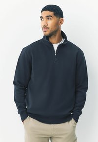 Marineblauwe half-zip sweatshirt met hoge kraag, zachte stof en geribbelde manchetten. Draagt over een wit T-shirt, gecombineerd met lichtbeige broek.
