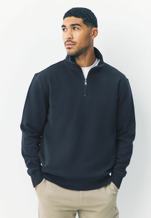 Marinblå halvmanschett sweatshirt med hög krage, mjukt tyg och ribbade muddar. Bärs över en vit T-shirt och matchas med ljus beige byxor.