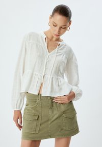 Blusa bianca textured con maniche lunghe e chiusura a bottoni, abbinata a una mini gonna in denim verde con tasche anteriori a patta.