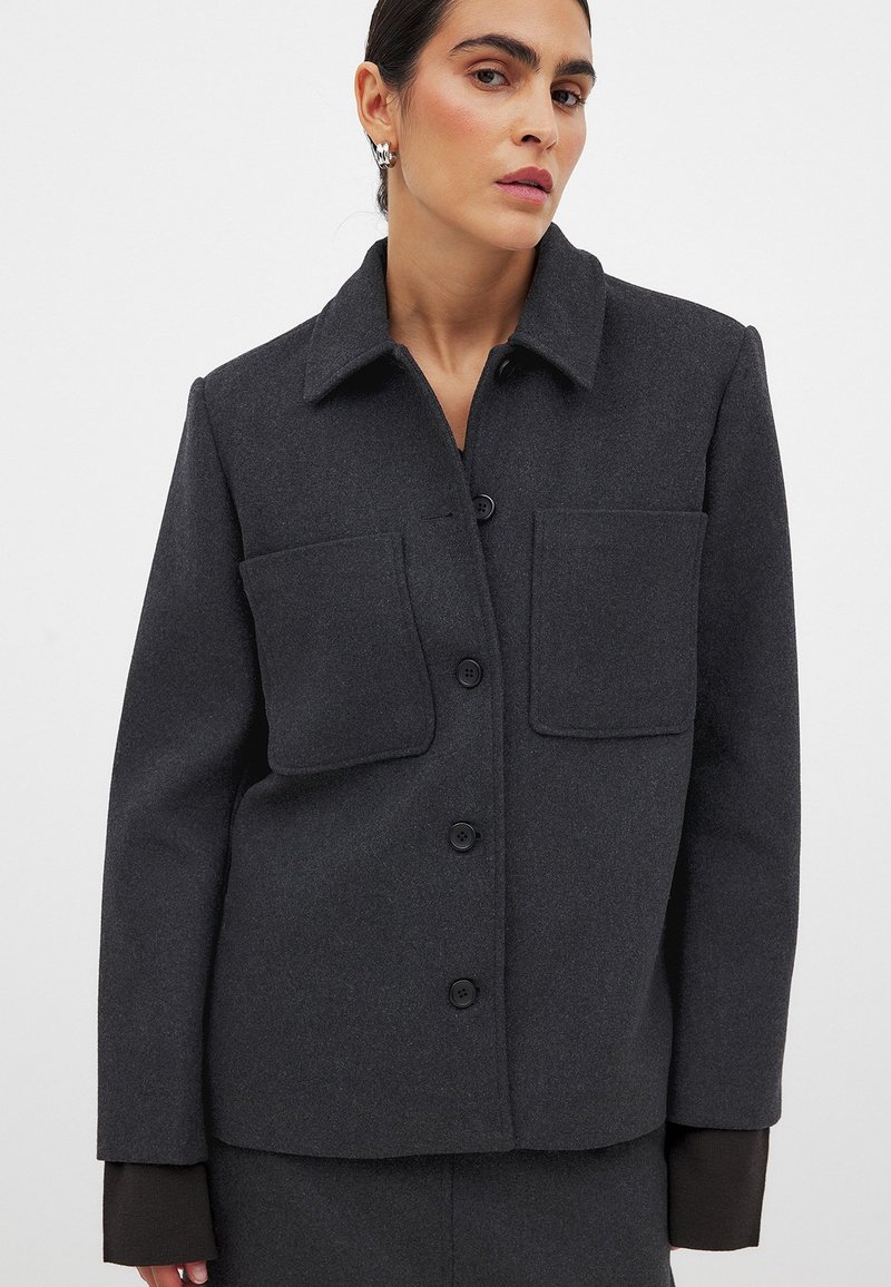 Temno siva jakna iz volnenega blend z ovratnikom, sprednjim gumbom za zapiranje in dvema velikima žepoma na prsih. Gladka tekstura.