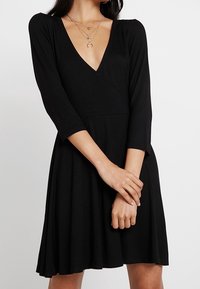 Svart omlottklänning med djup V-ringning, trekvartslånga ärmar och en utställd kjol. Tillverkad av mjukt, stretchigt material med en slät yta.