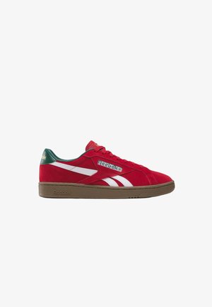 Zapatilla baja roja de Reebok con rayas blancas en los laterales, lengüeta verde en el talón y suela de goma marrón, vista desde el lado exterior.
