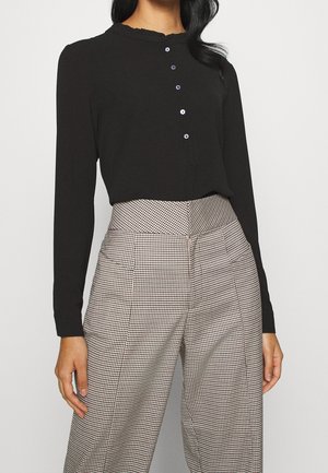 Kobieta w czarnej bluzce z długim rękawem, z małymi guzikami oraz w beżowo-czarnych, szerokich spodniach w kratę z wysokim stanem.