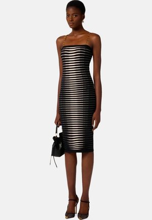 Donna che indossa un vestito midi aderente a righe nere e beige con spalline a catena dorata, tiene una piccola borsa nera e indossa tacchi alti neri.