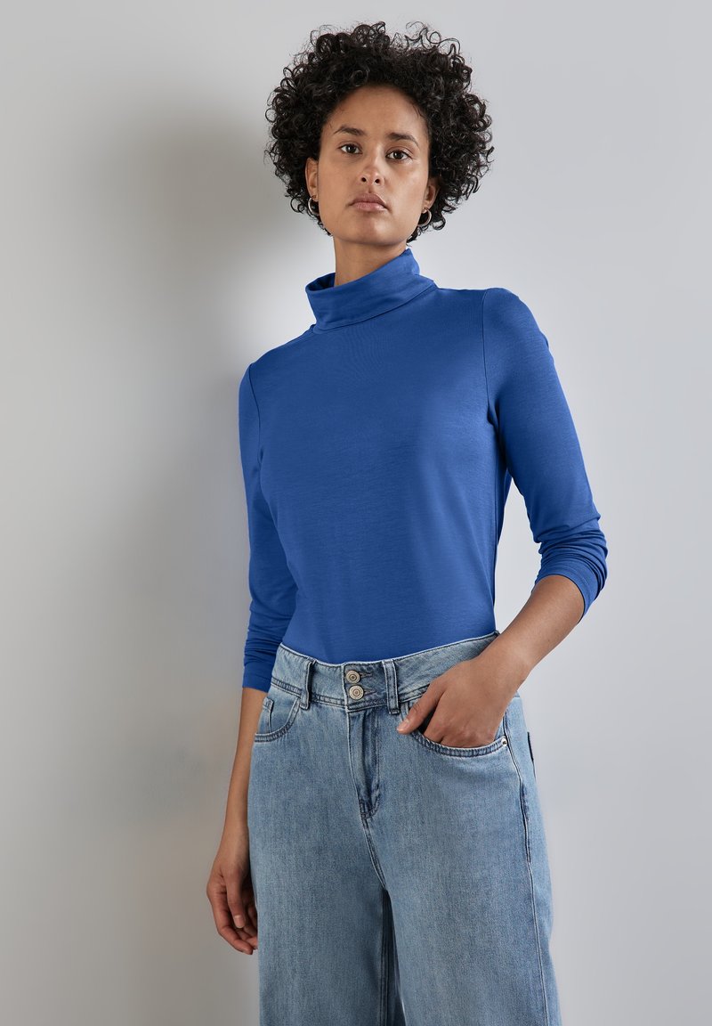 Blaues Rollkragenoberteil mit langen Ärmeln aus glattem Stoff, kombiniert mit hellblauen, hoch taillierten Denim-Jeans mit Knopfverschluss und Taschen.