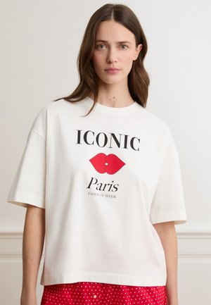 Frau mit langen braunen Haaren, die ein weißes übergroßes T-Shirt mit dem Schriftzug "ICONIC Paris Fashion Week" und einer Grafik roter Lippen trägt, kombiniert mit roten Punkten auf der Unterbekleidung.