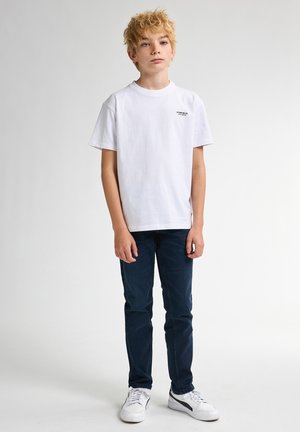 Camiseta blanca de algodón con mangas cortas y un pequeño logo negro en el pecho, combinada con unos vaqueros azul oscuro y zapatillas blancas.