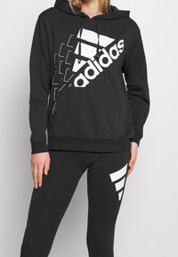 Sweat à capuche noir avec grand logo Adidas blanc et éléments de design ; associé à des leggings noirs avec un accent graphique blanc.