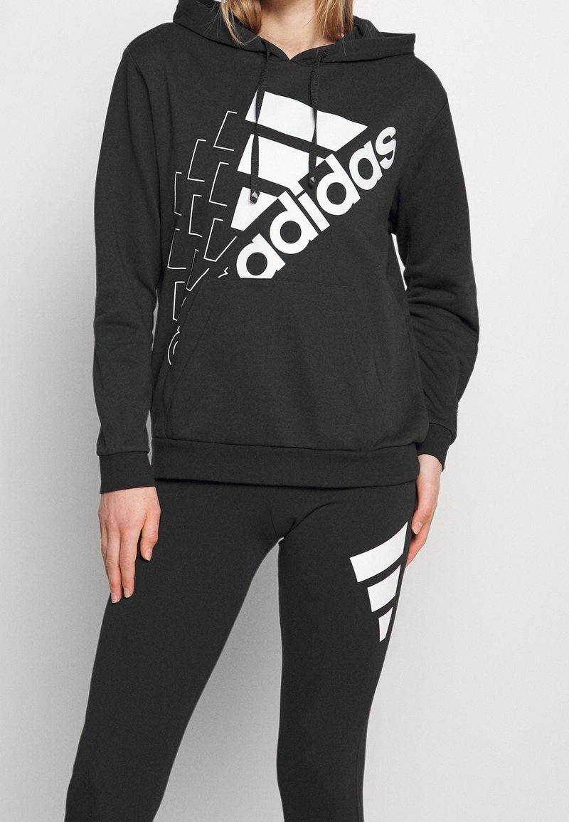 Sweat à capuche noir avec grand logo Adidas blanc et éléments de design ; associé à des leggings noirs avec un accent graphique blanc.