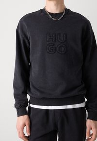 Sudadera de algodón negra con un logo en relieve "HUGO", puños y dobladillo acanalados, con un acabado texturizado y detalles de desgaste sutiles.