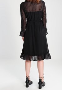 Černé šaty s puntíky vyrobené z průhledné látky, s dlouhými rukávy, elastickým páskem v pase a rozšířeným lemem s volánovými detaily.