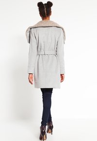 Cappotto in lana grigio con un ampio, morbido colletto in pelliccia sintetica, vita con cintura, maniche lunghe e design di lunghezza media. Indossato sopra jeans scuri e stivali con tacco.